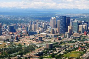 Aerial view of downtown Calgary with the Bow River, modern skyscrapers, and the Rocky Mountains visible in the distance.
